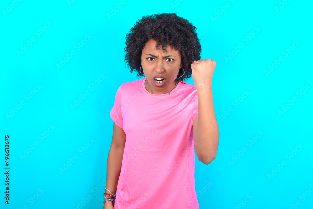 young girl with afro hairstyle wearing pink T-shirt over blue background angry and mad raising fist frustrated and furious while shouting with anger. Rage and aggressive concept.