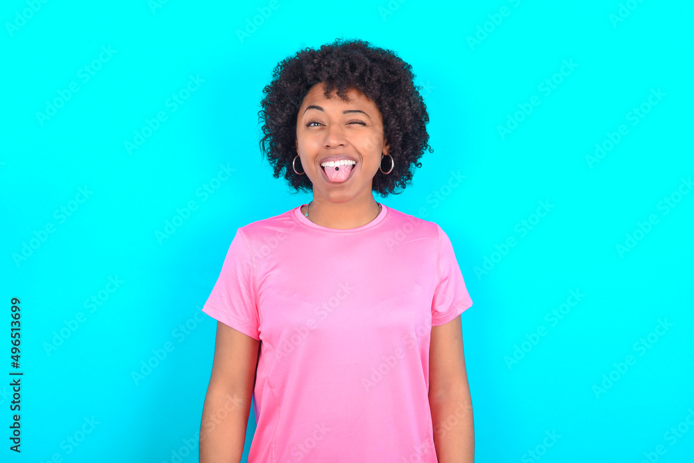 young girl with afro hairstyle wearing pink T-shirt over blue background sticking tongue out happy with funny expression. Emotion concept.