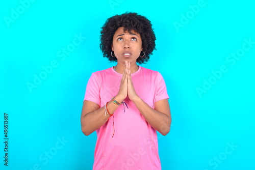 young girl with afro hairstyle wearing pink T-shirt over blue background begging and praying with hands together with hope expression on face very emotional and worried. Please God