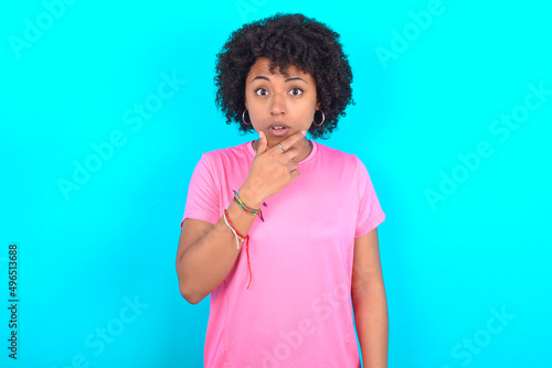young girl with afro hairstyle wearing pink T-shirt over blue background Looking fascinated with disbelief, surprise and amazed expression with hands on chin