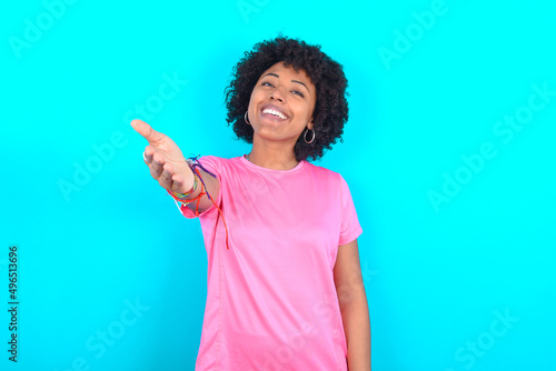 young girl with afro hairstyle wearing pink T-shirt over blue background smiling friendly offering handshake as greeting and welcoming. Successful business.