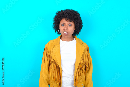Portrait of dissatisfied Young woman with afro hairstyle wearing yellow fringe jacket over blue smirks face, purses lips and looks with annoyance at camera, discontent hearing something unpleasant