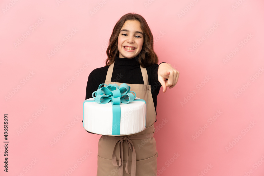 Little Pastry chef holding a big cake isolated on pink background surprised and pointing front Little Pastry chef holding a big cake isolated on pink background surprised and pointing front