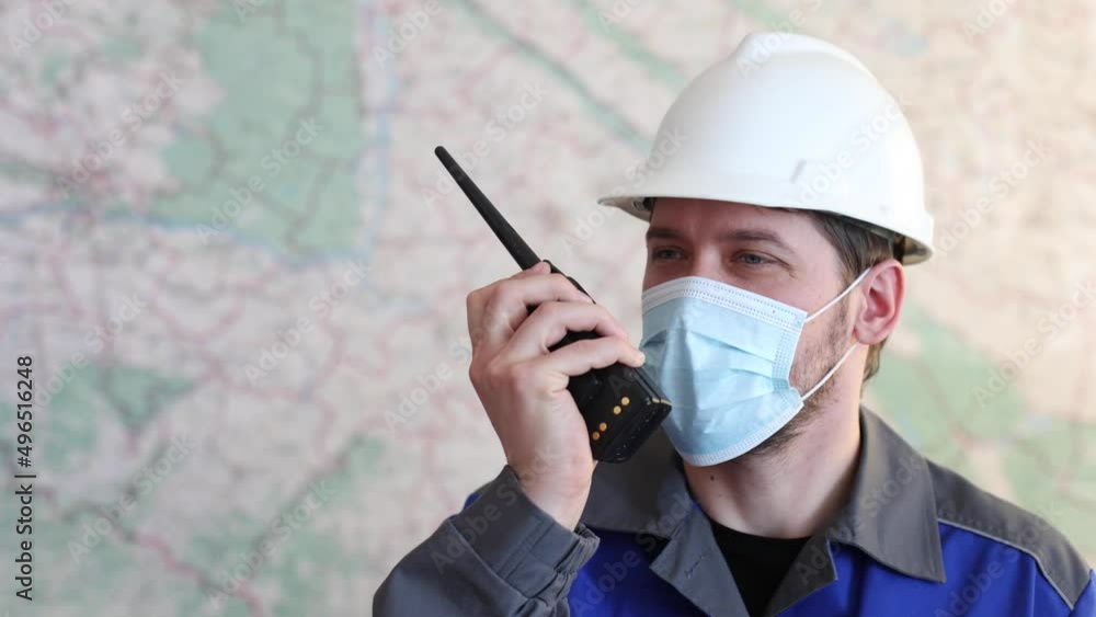 Positive worker in a helmet, uniform and mask uses a walkie-talkie against the background of a geographical map. Radio engineer gives instructions remotely and uses a mask to protect against viruses Positive worker in a helmet, uniform and mask uses a walkie-talkie against the background of a geographical map. Radio engineer gives instructions remotely and uses a mask to protect against viruses