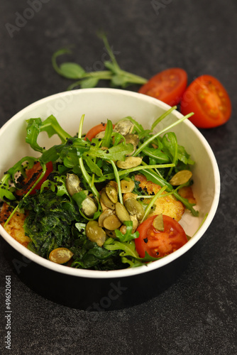 Vegetarian salad with potato, tomatoes, arugula and 
pumpkin seeds in round paper take-away container on dark background. Healthy food delivery concept. Free space for text.