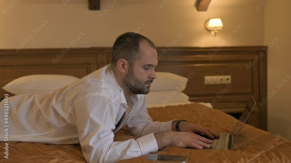 Young businessman working at a laptop lying on a bed in a hotel room. Confident male working remotely after arriving on business trip.