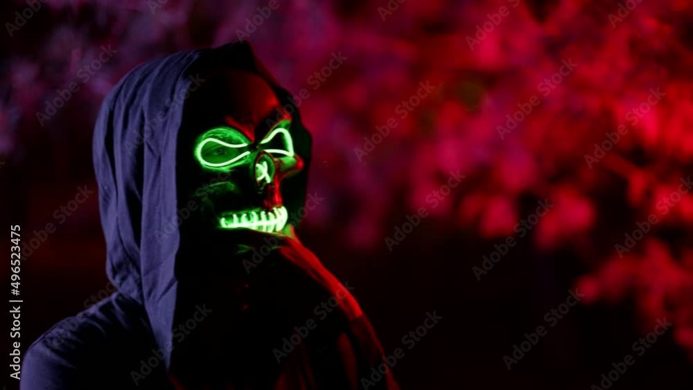 portrait of a person in a glowing mask of death and in a hood against a background of branches illuminated by red light. the character rubs chin thoughtfully and looks around. the dark key portrait of a person in a glowing mask of death and in a hood against a background of branches illuminated by red light. the character rubs chin thoughtfully and looks around. the dark key