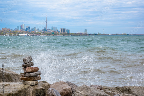 A small inukshuk (pile of rocks) is seen in the foreground, being splashed by the chilly water of Lake Ontario, with the Toronto city skyline in the background on an overcast day. photo