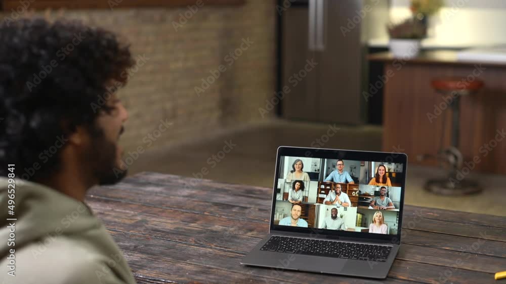 Young guy relaxed sitting at the table and talking online via video call with group of friends or employees. Man using app on laptop for video meeting with many people together Young guy relaxed sitting at the table and talking online via video call with group of friends or employees. Man using app on laptop for video meeting with many people together