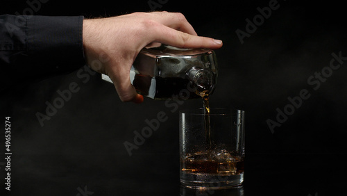 Hand of barman pouring golden whiskey, cognac or brandy from bottle into glass with ice cubes on black background. Bartender pouring glass of alcohol drink bourbon, rum. Copy-space