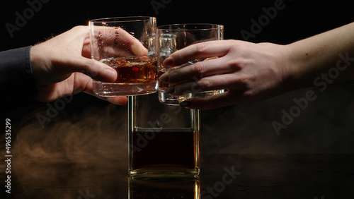 Two hands with glasses of cognac whiskey with ice cubes making cheers on black background. Celebration of business success, Christmas, anniversary, birthday. Raising toast with glasses of bourbon, rum