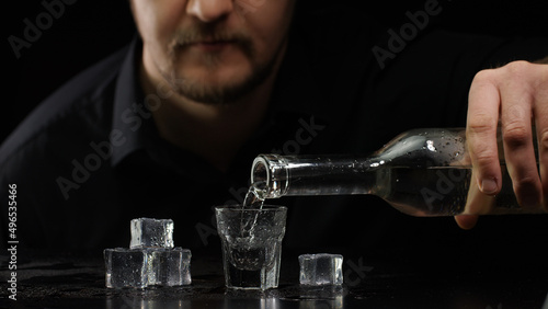 фотографія Bartender man pouring up frozen vodka or sake from bottle into shot glass with ice cubes on black background