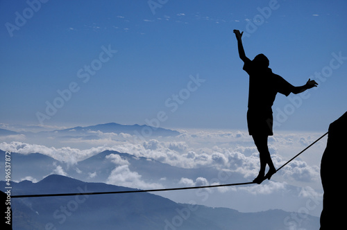 Silhouette of young man balancing on slackline high above clouds and mountains. Slackliner balancing on tightrope beautiful colorful sky and clouds behind, highline silhouette.
