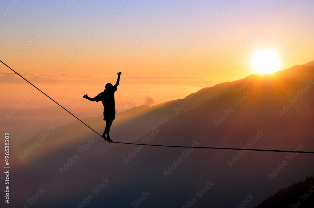 © vp3k - Silhouette of young man balancing on slackline high above clouds and mountains. Slackliner balancing on tightrope during sunset, highline silhouette. © vp3k - Silhouette of young man balancing on slackline high above clouds and mountains. Slackliner balancing on tightrope during sunset, highline silhouette.