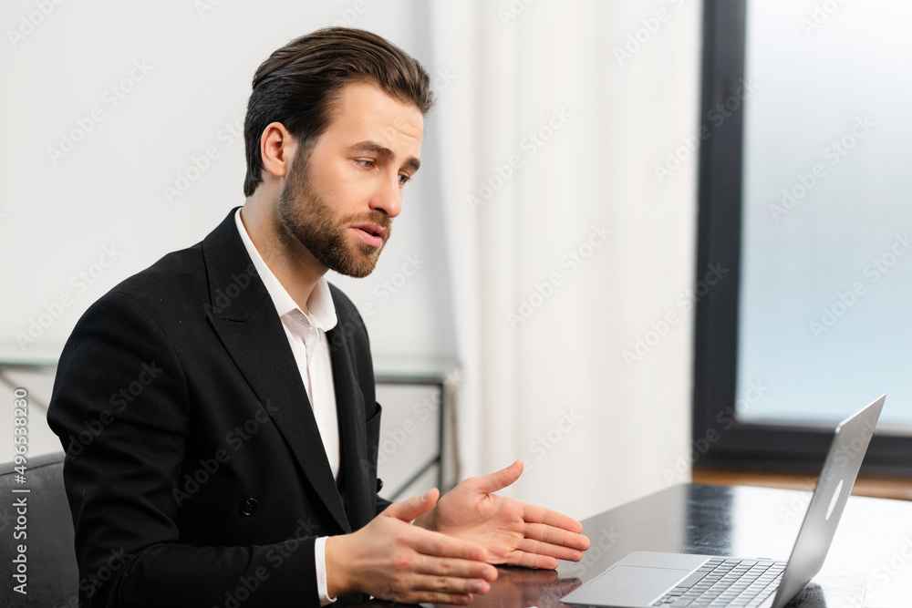 Side view of caucasian office employee talking via video connection while sitting at the desk. A guy using laptop for video call in contemporary office space Side view of caucasian office employee talking via video connection while sitting at the desk. A guy using laptop for video call in contemporary office space