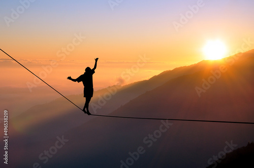 Silhouette of young man balancing on slackline high above clouds and mountains. Slackliner balancing on tightrope during sunset, highline silhouette. photo