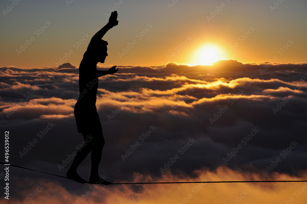 Silhouette of young man balancing on slackline high above clouds and mountains. Slackliner balancing on tightrope during sunset, highline silhouette. Silhouette of young man balancing on slackline high above clouds and mountains. Slackliner balancing on tightrope during sunset, highline silhouette.