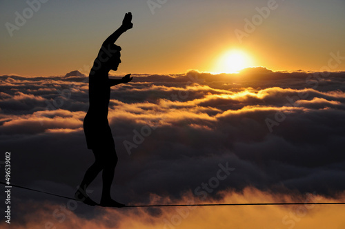 Silhouette of young man balancing on slackline high above clouds and mountains. Slackliner balancing on tightrope during sunset, highline silhouette.