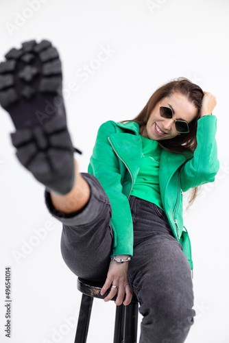 Portrait of smiling lady in casual clothes sitting on chair with raised leg. Attractive woman with trendy accessories and stylish wear closeup. Fashion photo, posing on white background in studio