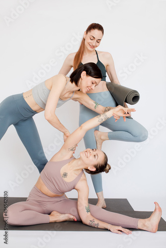 three beautiful girls smile and practice yoga. Yoga teacher helps to stretch on the mat in a pose on a white background.
girlfriends are dressed in sports leggings and tops. Long shote