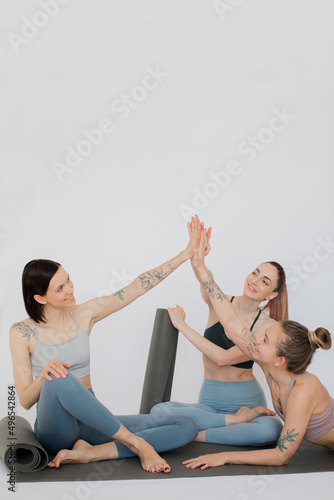 three beautiful girls smile, relax sitting on a yoga mat, make a handshake, joyfully give five in a white studio.
models are dressed in sports leggings and tops
