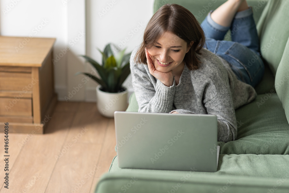 Young beautiful smiling woman using laptop lying on the couch. Concept of freelance job, home office. Female using technology for learning, shopping, e-commerce, communication, social media