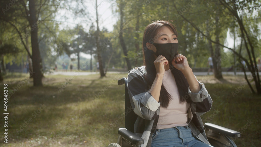 A beautiful disabled asian lady with long dark hair is putting a protective mask on her face in a public park A beautiful disabled asian lady with long dark hair is putting a protective mask on her face in a public park
