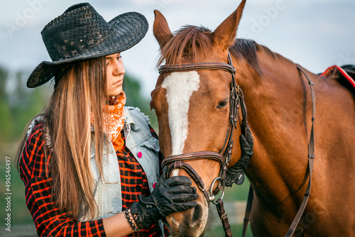 A cowgirl stands near her horse in a field at sunset.