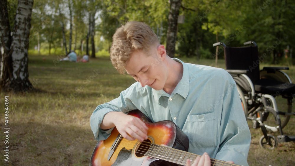 Fototapeta premium A young disabled blonde man is sitting on the plaid in the public park and playing guitar professionally