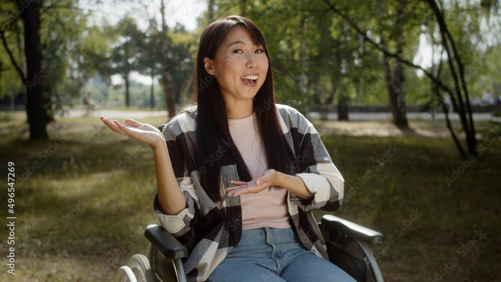 An attractive cheerful asian girl with long dark hair is talking to the camera, smiling and gesturing, sitting in the wheelchair in the public park An attractive cheerful asian girl with long dark hair is talking to the camera, smiling and gesturing, sitting in the wheelchair in the public park