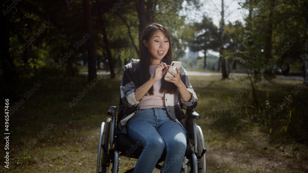 A pretty girl is sitting in a wheelchair in a public park and looking into the screen of her mobile phone, searching through the Internet and smiling