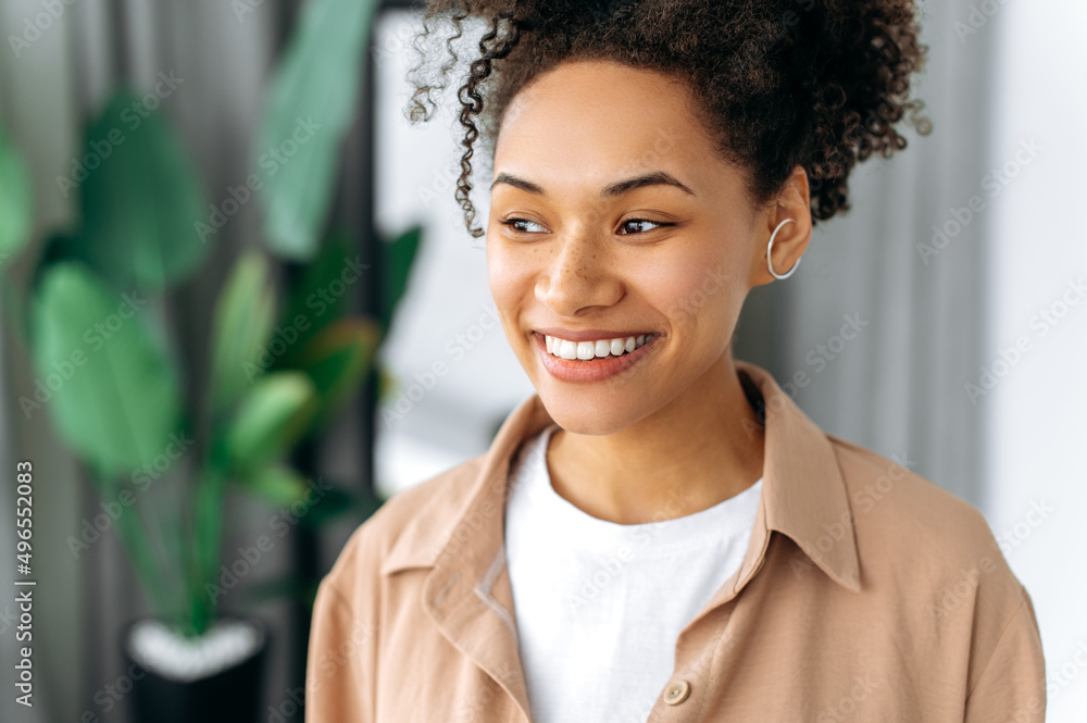 Beautiful joyful positive millennial African American girl with curly hair and freckles, wearing casual clothes, standing at home, looking away, smiling happily Beautiful joyful positive millennial African American girl with curly hair and freckles, wearing casual clothes, standing at home, looking away, smiling happily