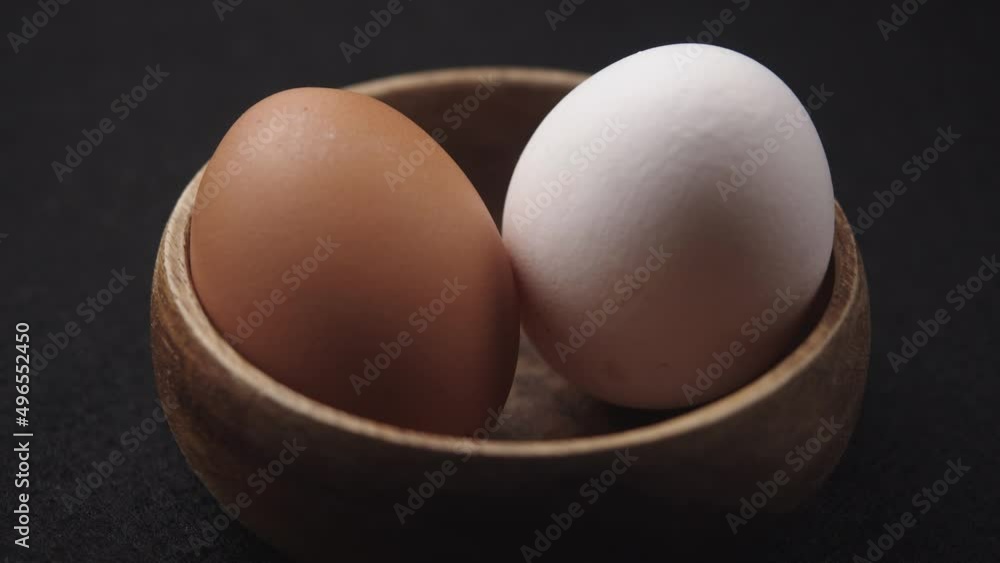 White and Brown Eggs in wooden bowl on black background. Close-up of two fresh, organic and healthy chicken egg rotate. White, Brown Eggs are rolling on the clean black table. Easter eggs background. White and Brown Eggs in wooden bowl on black background. Close-up of two fresh, organic and healthy chicken egg rotate. White, Brown Eggs are rolling on the clean black table. Easter eggs background.