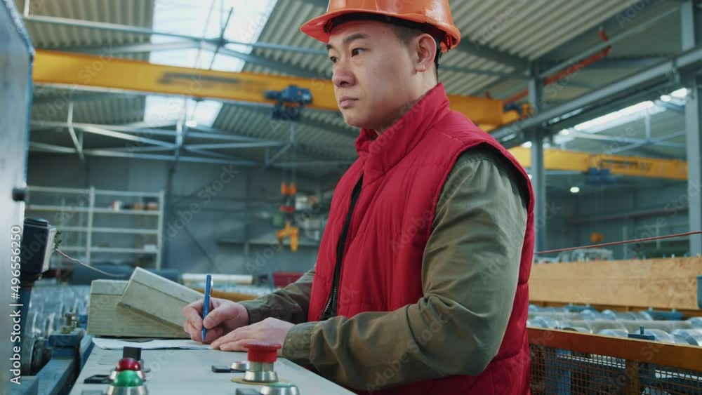 Asian engineer man wearing safety uniform and hard hat stand checks and writes on paper. Production processes in factory. Look around. Inspector in warehouse. Asian engineer man wearing safety uniform and hard hat stand checks and writes on paper. Production processes in factory. Look around. Inspector in warehouse.