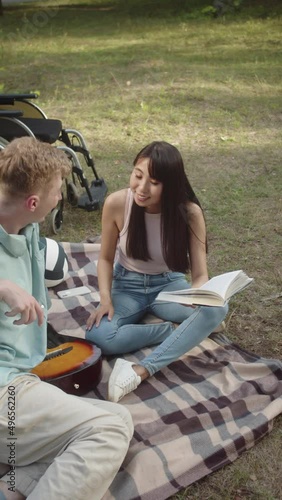 Sitting on the plaid in a public park, a young asian girl with long dark hair is reading a book for her disabled friend in a blue shirt