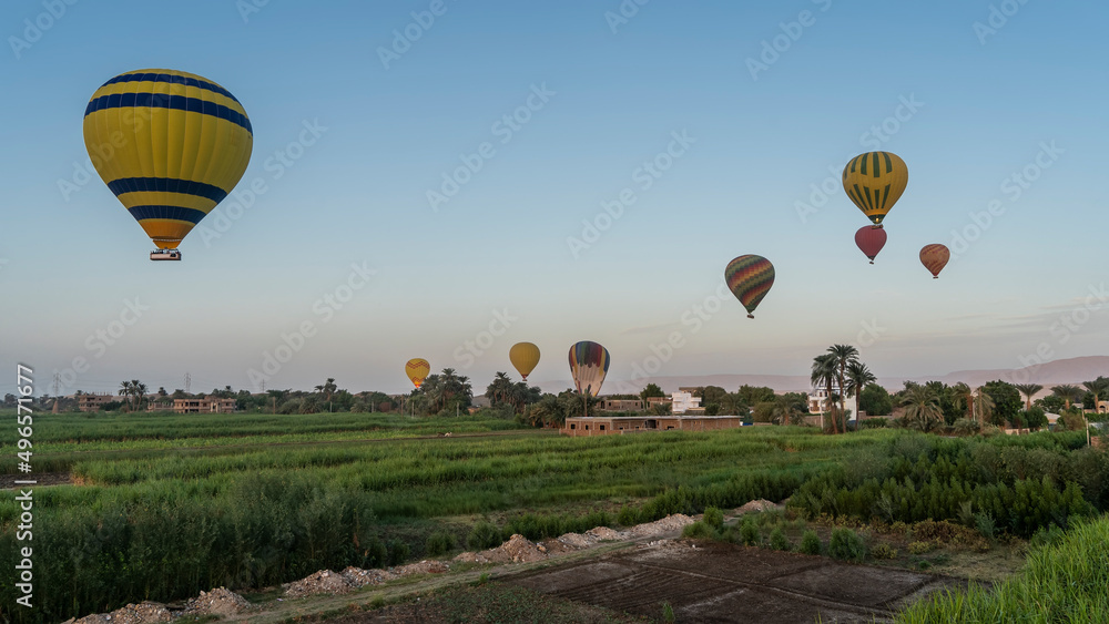 Bright balloons are flying over sugar cane plantations. Farmhouses are visible in the distance. Sand dunes against a blue sky. Egypt. Luxor