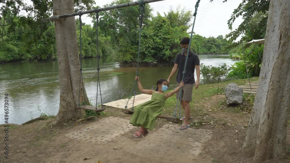 Asian child or kid girl wearing face mask playing ride on swing and happy fun with father or family on holiday relax travel at river playground or public park on nature tree garden and happiness sound Asian child or kid girl wearing face mask playing ride on swing and happy fun with father or family on holiday relax travel at river playground or public park on nature tree garden and happiness sound