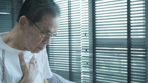 senior asian man coughing wearing glasses, after waking up in the morning, in the bedroom, wearing a white shirt, covering his mouth with hands photo