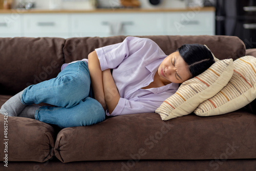Young Asian woman suffering from abdominal pain at home lying on the couch at home. Female suffering from severe spasms holding her stomach © kucherav