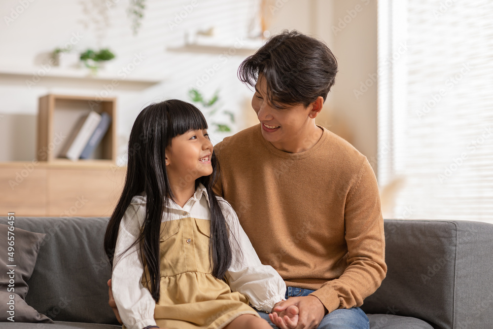 Smiling Asian father embracing daughter sitting on couch, posing for family photo indoors. Happy young dad with little child girl, resting on sofa in the living room. Smiling Asian father embracing daughter sitting on couch, posing for family photo indoors. Happy young dad with little child girl, resting on sofa in the living room.
