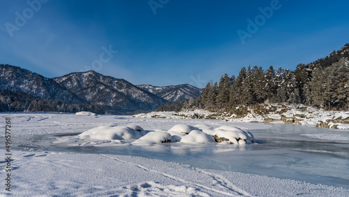 The frozen river is covered with snow. Picturesque boulders on turquoise ice. Mountain range and coniferous forest against the blue sky. Altai. Katun