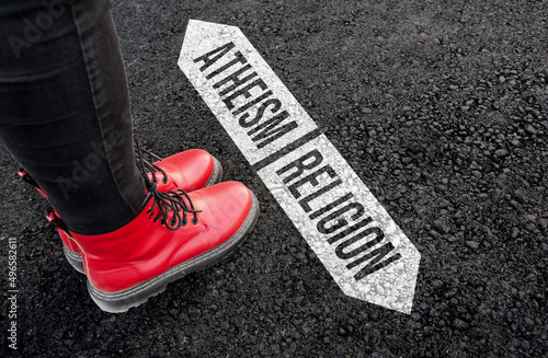 woman with boots are standing on asphalt next to arrow signs of words atheism and religion