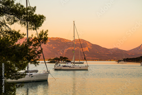 View on sailing yachts staying on anchor in the Russian bay  half island Poros  Greece with small island and half island Methana in the background and beautiful sunset lights