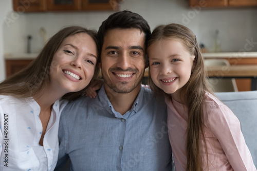 Happy young couple of parents and sweet daughter child head shot portrait. Mom, dad, kid hugging, relaxing together at home, looking at camera with toothy smiles. Parenthood concept