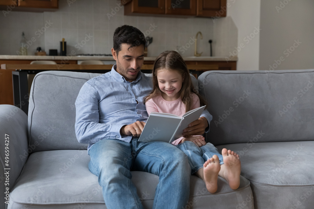 Loving caring dad and cute preschool daughter girl reading book on couch together, hugging, sitting on sofa, enjoying leisure at home together. Handsome father telling fairy tale to cheerful kid Loving caring dad and cute preschool daughter girl reading book on couch together, hugging, sitting on sofa, enjoying leisure at home together. Handsome father telling fairy tale to cheerful kid