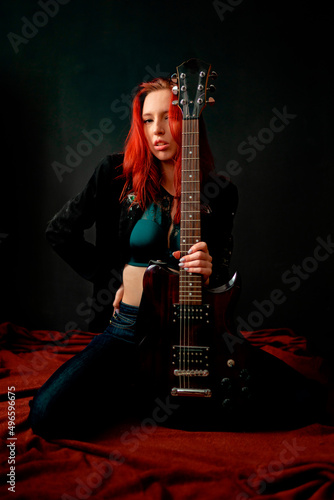 Sexy and brutal rocker punk or metal red-hair girl with an electric guitar in a black jacket and jeans is sitting on the floor on her knees in a defiant pose © evgenzz