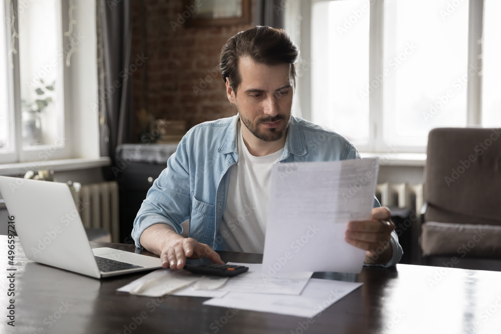Focused millennial man paying domestic bills, using calculator, accounting app on laptop, reading paper document, invoice, bank notice. Entrepreneur, businessman doing financial paperwork Focused millennial man paying domestic bills, using calculator, accounting app on laptop, reading paper document, invoice, bank notice. Entrepreneur, businessman doing financial paperwork
