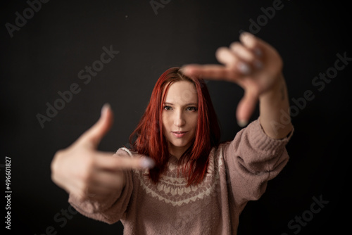 a beautiful and natural smiling red-haired skinny girl in a knitted sweater makes an imaginary lens or camera frame gesture with her fingers and looks through it at the viewer