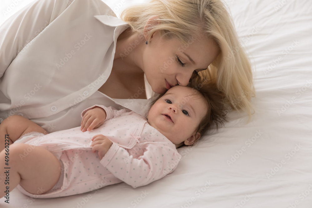 Happy loving mom kissing sweet few month baby with care, affection, tenderness, resting on bed mattress with white sheet. New mother enjoying leisure time with little infant kid in bedroom Happy loving mom kissing sweet few month baby with care, affection, tenderness, resting on bed mattress with white sheet. New mother enjoying leisure time with little infant kid in bedroom
