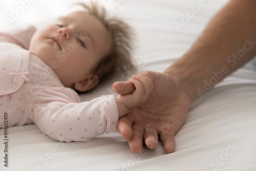 Serene adorable sleepy baby catching dads thumb, holding finger, lying on back, sleeping on bed. Cute infant child in pink bodysuit sleeping under parent supervision. Close up of hand, cropped shot